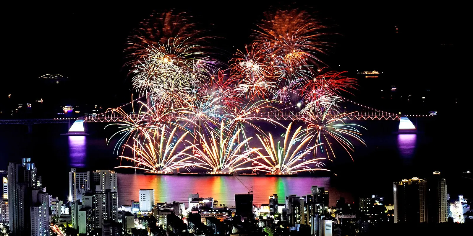 広安大橋の上で花火が一斉に開き、手前の広安里の高層ビルと水面に光が映っている様子