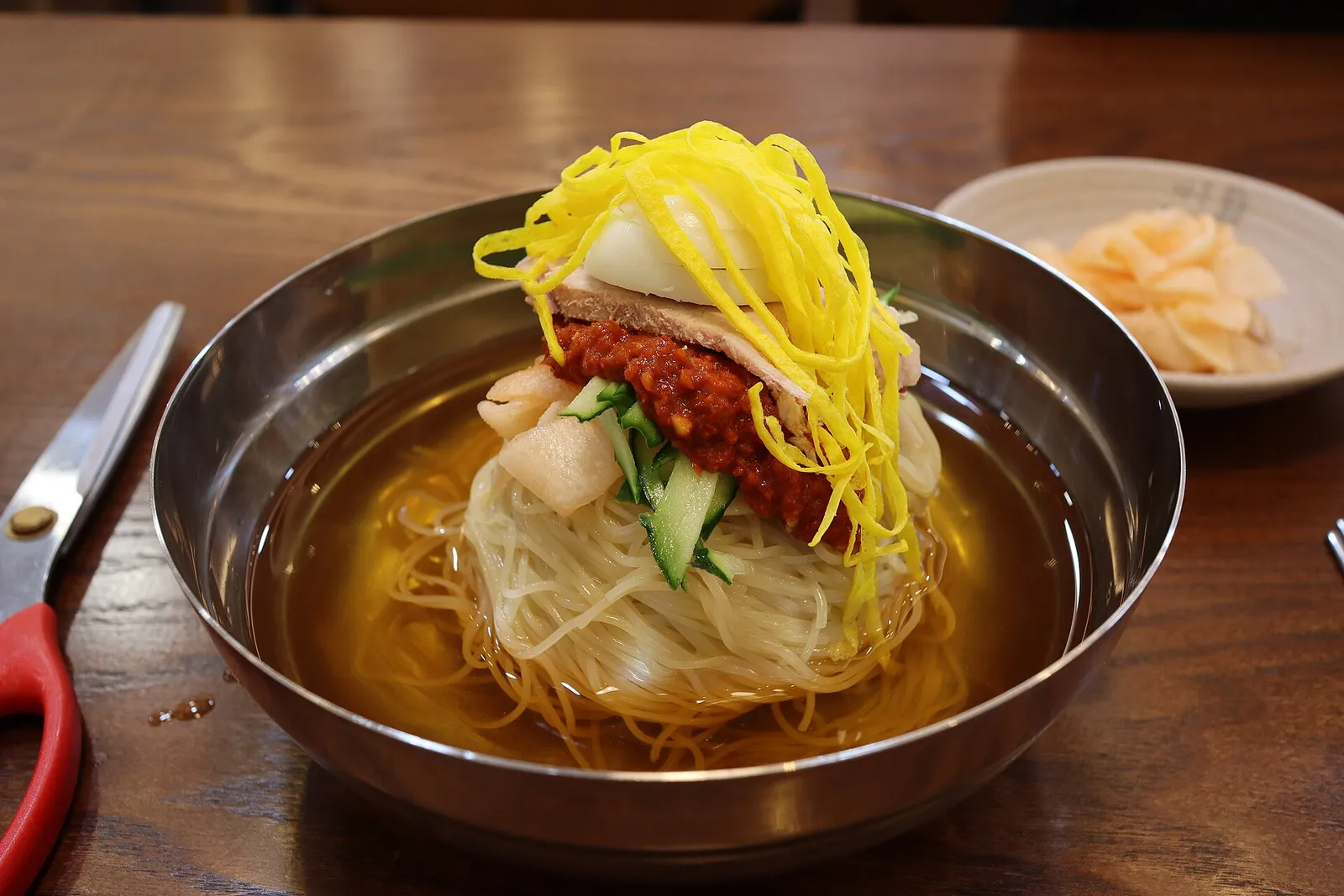 A steel bowl of milmyeon, chewy wheat noodles in cold broth topped with red sauce and egg