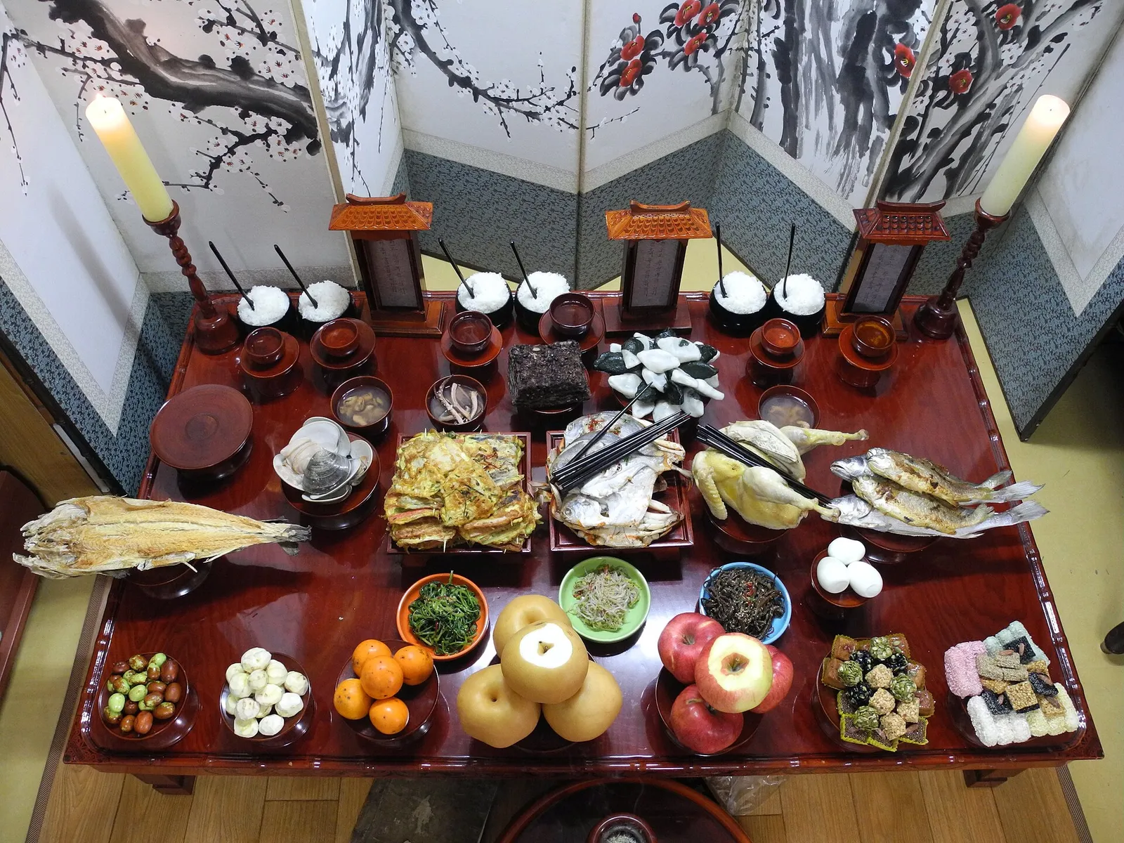 A charye ancestral rite table set before a folding screen, with rice and soup, pan-fried dishes and fish, and pears and apples stacked in tiers