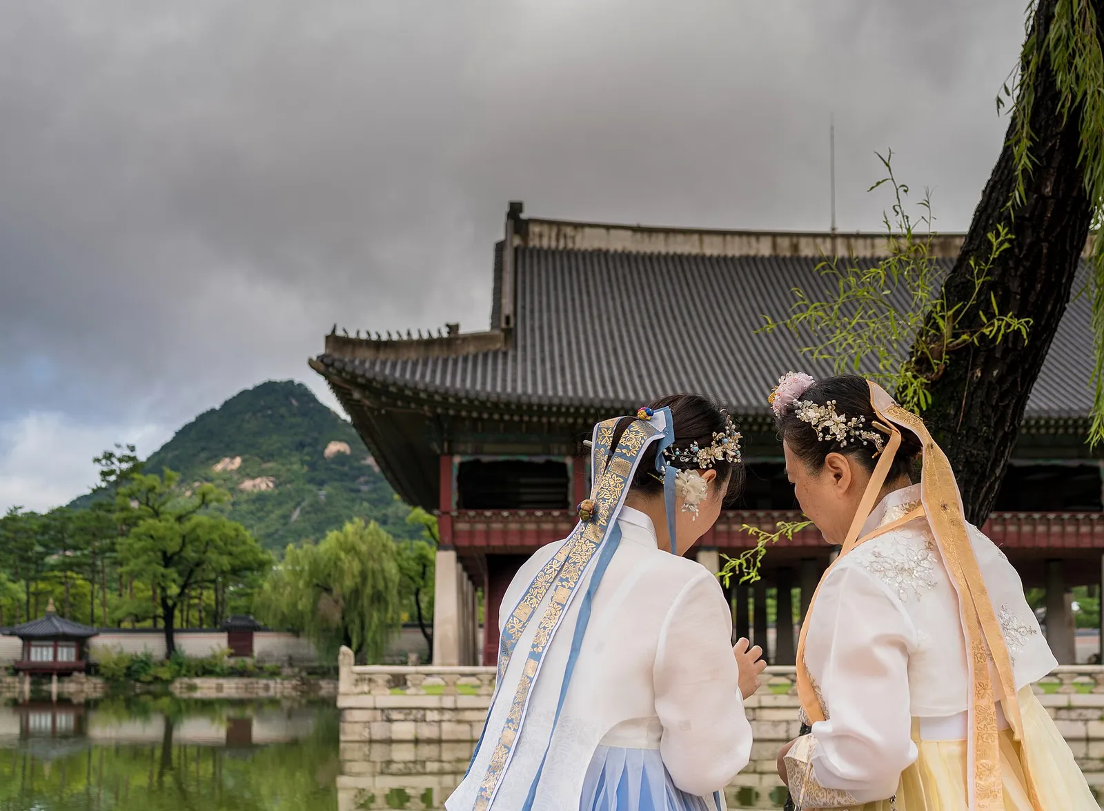 Two people in hanbok standing in front of Gyeonghoeru pavilion at Gyeongbokgung, with the pond and Inwangsan behind them