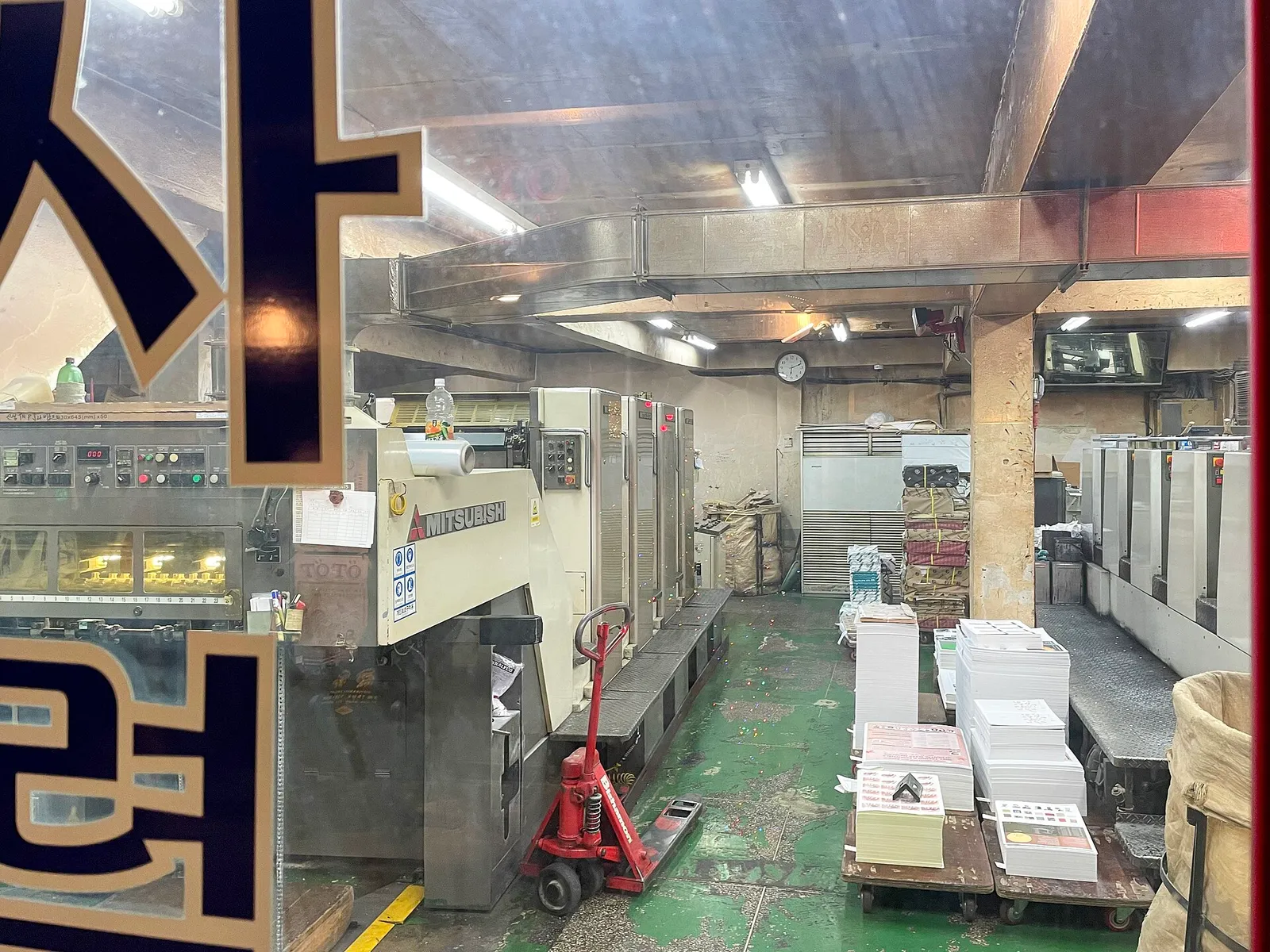 Inside a print shop seen through the window, printed sheets stacked beside a Mitsubishi press