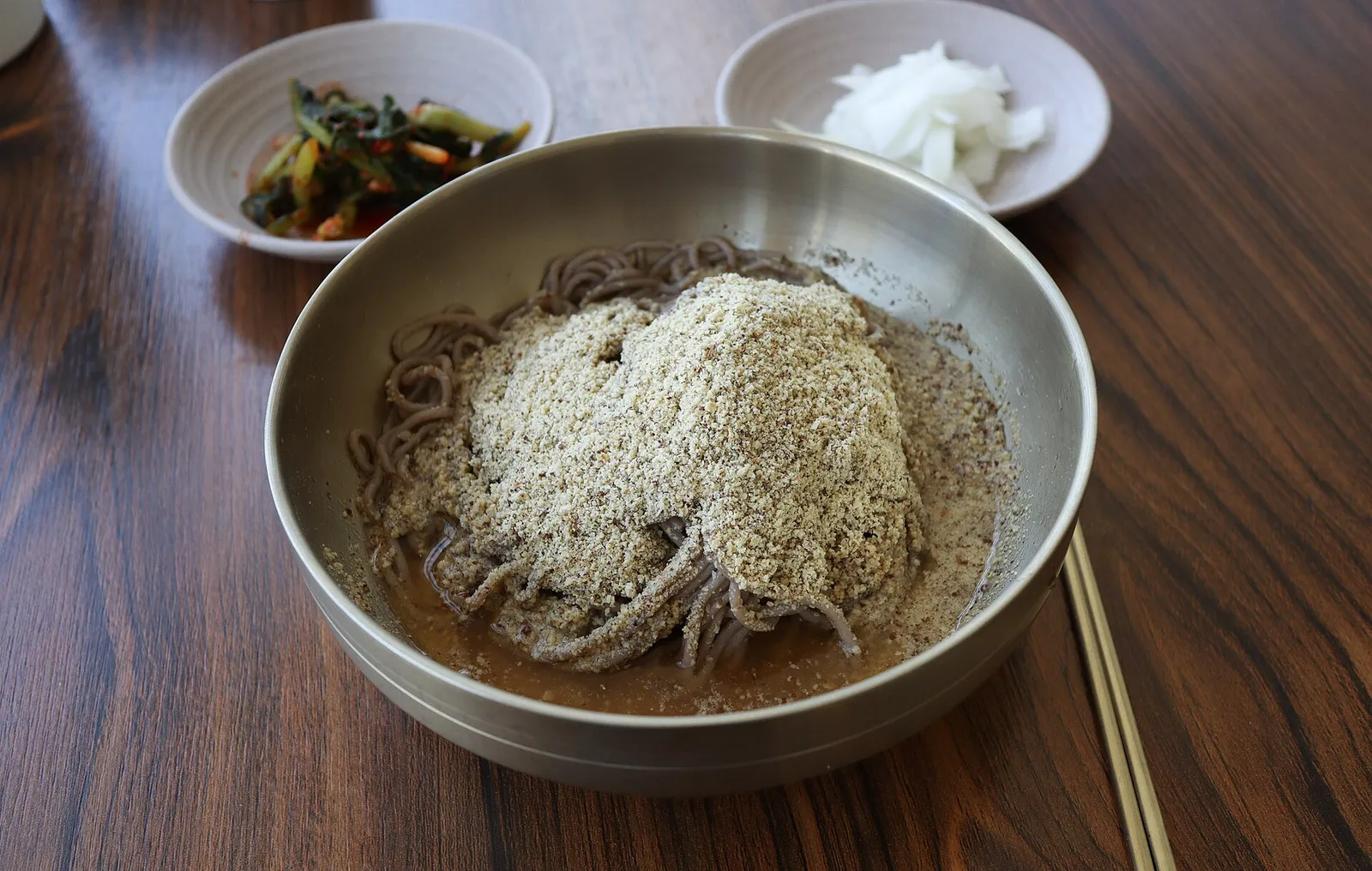 ステンレスの器に盛られたマッククス、そば粉の麺の上にすりエゴマがたっぷりのり、小皿のおかずが並ぶ