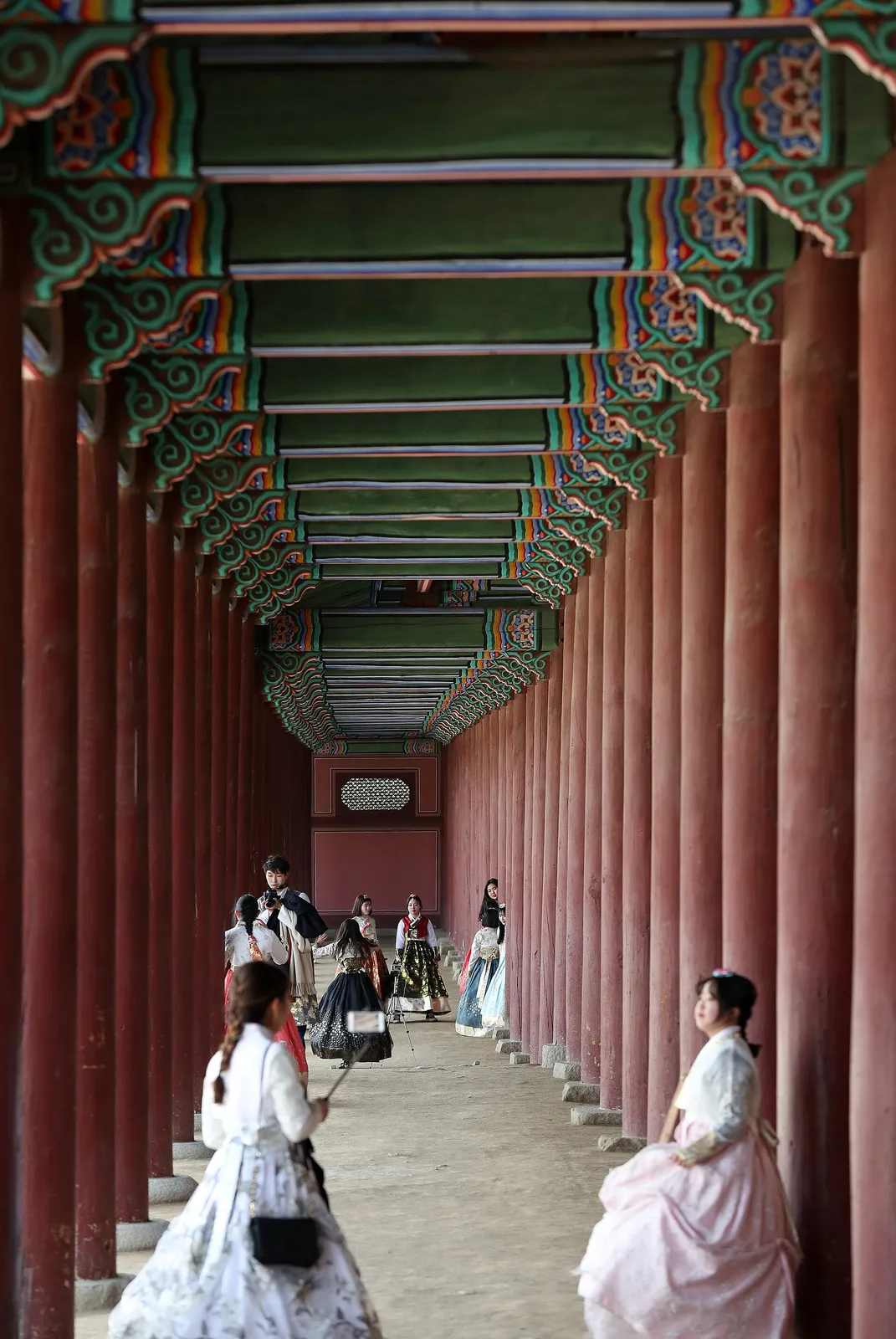 Visitors in hanbok standing between the red pillars of a Gyeongbokgung colonnade, with someone shooting from a tripod further in