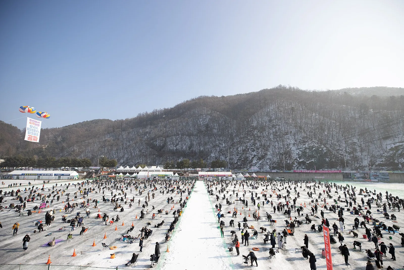 凍った華川川を埋め尽くす氷釣りの人波、奥に雪をかぶった山と「Sancheoneo Ice Fishing」の英語看板