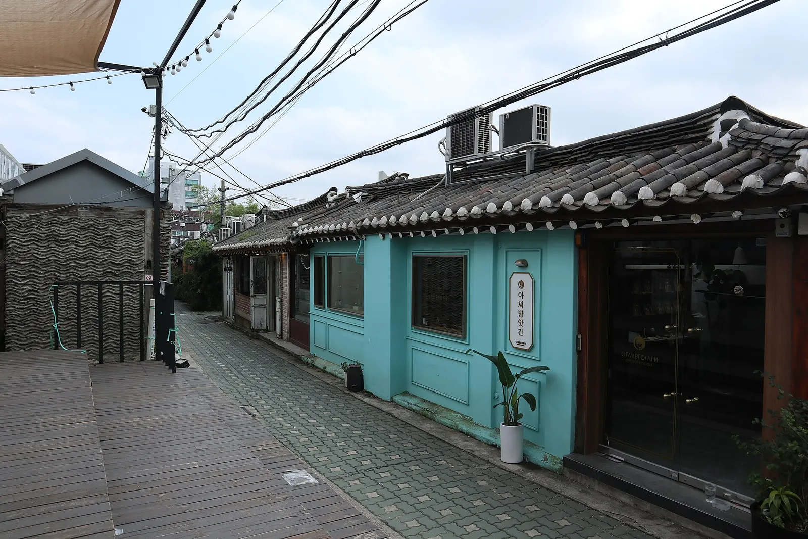 An Ikseon-dong alley of low continuous hanok rooflines, with a wooden deck laid in front of a mint-green shop
