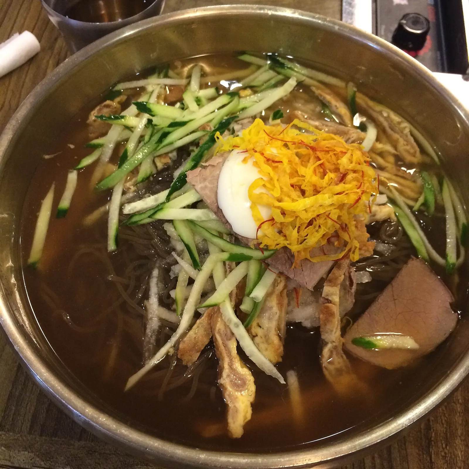 真鍮の器に盛られた晋州の水冷麺。細切りのきゅうりと卵、黄色い錦糸卵、薄く焼いたユクチョンがのっている