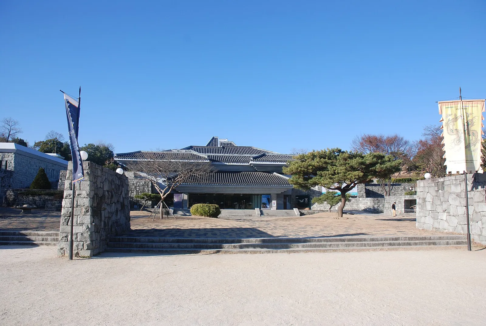 瓦屋根をのせた国立晋州博物館の建物と前庭、石垣と松の木