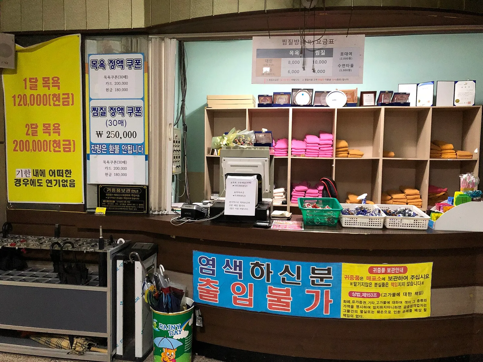 チムジルバンのカウンター、ピンクと黄色のタオルが棚ごとに積まれ、その上に料金表とクーポンの案内が貼ってある