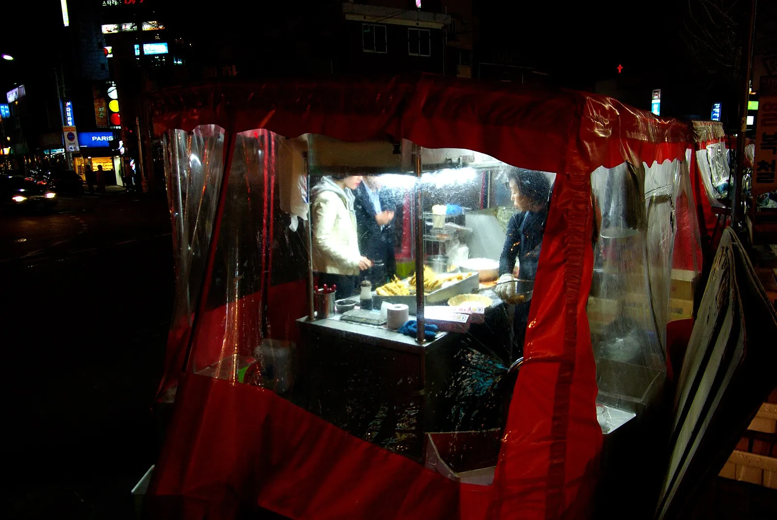 A red-canopied street tent bar at night, cooking counter and customers visible through the plastic sheeting