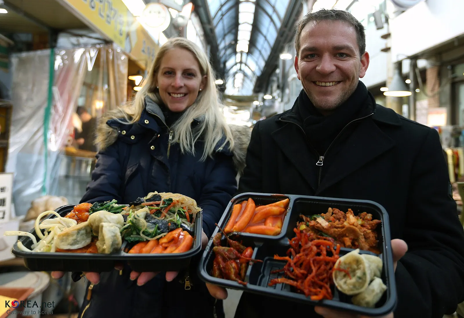 Two people in a market passageway holding up black lunchboxes with side dishes packed into the compartments